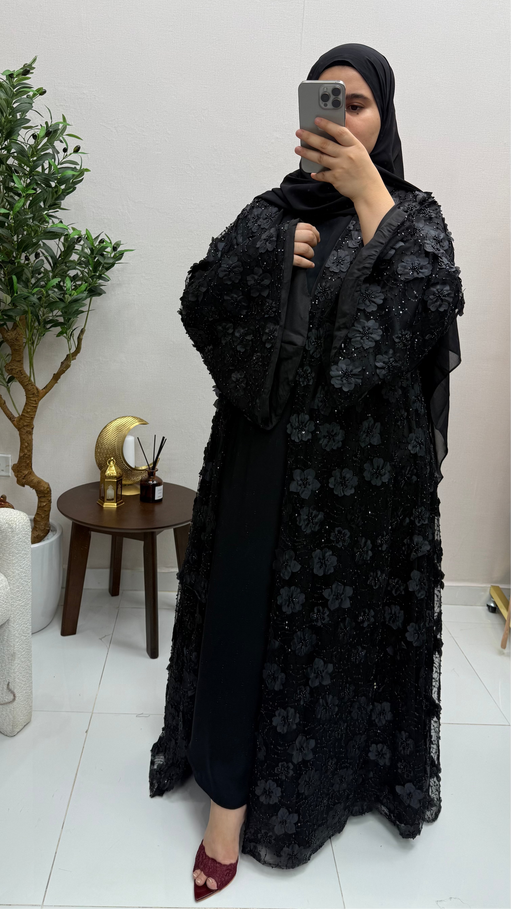 Woman taking a mirror selfie wearing a black floral abaya in a room with a plant and small table.