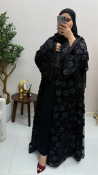 Woman taking a mirror selfie wearing a black floral abaya in a room with a plant and small table.